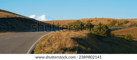 Winding mountain highway. A snowy volcano peak in the background. Shooting with a long lens. Copy space. Royalty-Free Stock Photo #2389802141