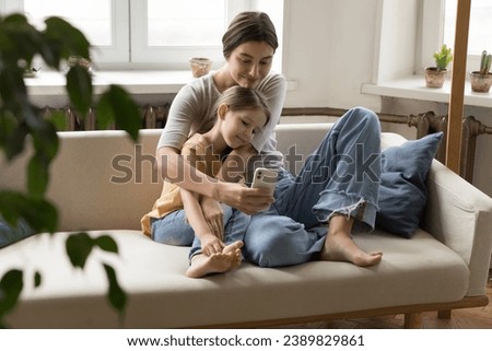 Loving young mother sit on sofa hug little girl spend free time at home, take selfie, watch funny video, use app. Happy family use social networks for entertainment. Internet, modern application usage