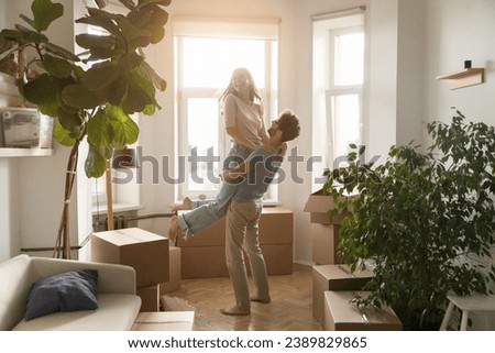 Happy husband lifts his young beautiful wife, laughing, swirling, enjoy move-in day to new, first house, big carton boxes nearby in unfurnished sunny living room. Bank loan for young family, new life Royalty-Free Stock Photo #2389829865