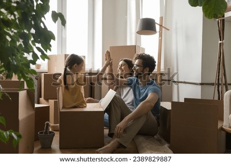 Young family with little daughter celebrate relocation day, feel happy, giving high five gesture seated on floor near heap of cardboard boxes. Bank loan, tenancy, first dwelling, housing improvement Royalty-Free Stock Photo #2389829887