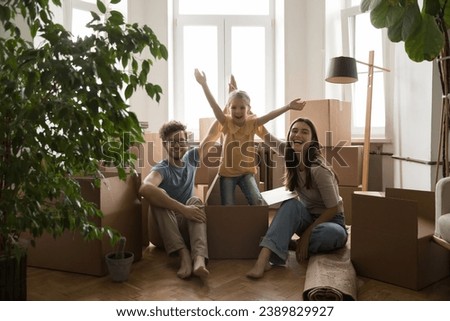 Little girl jumps out from cardboard box, having fun, enjoy relocation day posing smile look at camera with young parents, sit on floor near heap of cardboard boxes. Bank loan, new house, housewarming Royalty-Free Stock Photo #2389829927