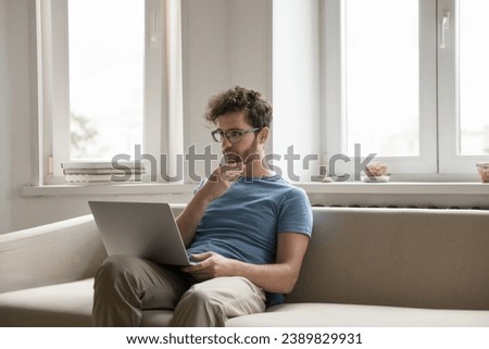Thoughtful man freelancer sitting with laptop on sofa work on project, thinking about difficult tasks touch chin, solve problem. Serious guy read message, looking for inspiration for new ideas at home Royalty-Free Stock Photo #2389829931