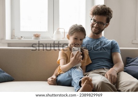 Happy young man sit on cozy couch with little daughter use mobile phone, communicate, play online game. Smiling cute girl enjoy weekend together with father, watch funny video. Modern tech, internet