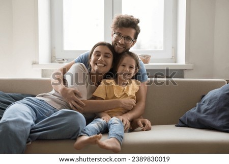 Loving young man hug wife and little daughter sitting on sofa, happy family smile pose looking at camera. Happy parents spend weekend time with cute kid at home, enjoy time together. Parenthood, love Royalty-Free Stock Photo #2389830019