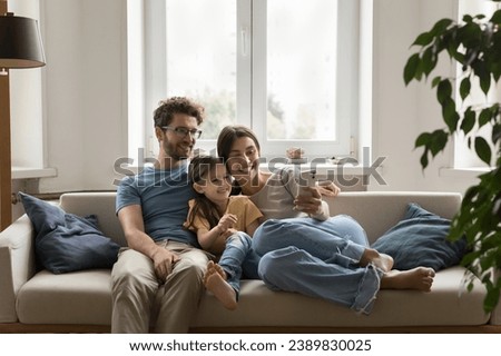 Smiling young spouses sit on sofa with little daughter hold smartphone, take self-portrait on camera. Family spend weekend time make video conference, using internet and videocall app on mobile phone Royalty-Free Stock Photo #2389830025