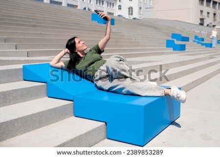 smiling brunette woman 40-49 years old takes a selfie, a photo of herself on her mobile phone, lying on a bench in the city on a sunny summer day