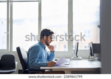 Busy serious young business man working on laptop computer holding documents at workplace. Latin professional businessman employee or manager thinking of financial management in office. Royalty-Free Stock Photo #2389861799