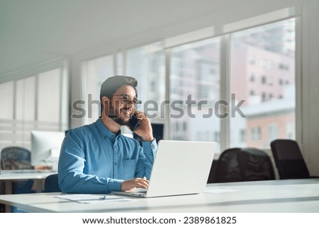 Young happy busy latin businessman talking on phone using laptop computer in office. Smiling hispanic business man making call on mobile cellphone, consulting client or having work cell conversation. Royalty-Free Stock Photo #2389861825