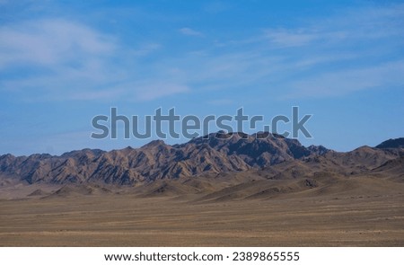 a rugged mountain range under a vast blue sky, with undulating hills in the foreground and sharp, rocky peaks in the distance creating a dramatic landscape Royalty-Free Stock Photo #2389865555