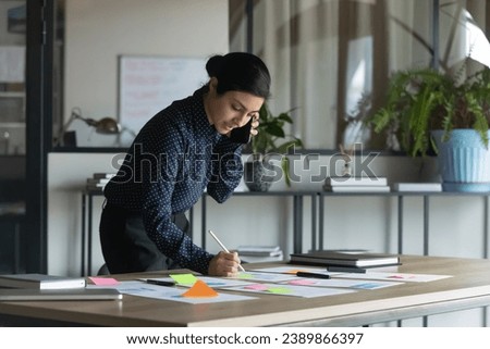 Serious busy multitasking young Indian businesswoman working with scrum arranged sticky papers, documents, graphic reports, writing notes, making call on cellphone, talking on mobile phone Royalty-Free Stock Photo #2389866397