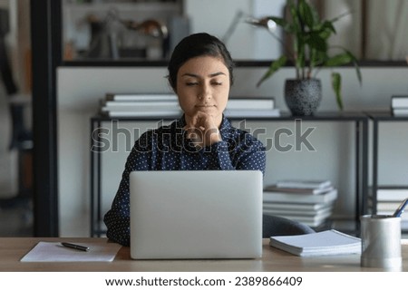 Serious Indian female student using laptop for studying, learning online, watching webinar, virtual business training, lecture. Young office employee working at computer, reading email message Royalty-Free Stock Photo #2389866409