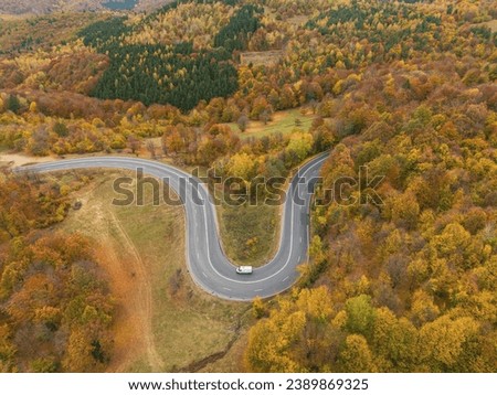 Aerial view of mountain road in beautiful forest at sunset in autumn. Top view from drone of winding road in woods. Colorful landscape with curved roadway, trees with orange leaves in fall. Travel