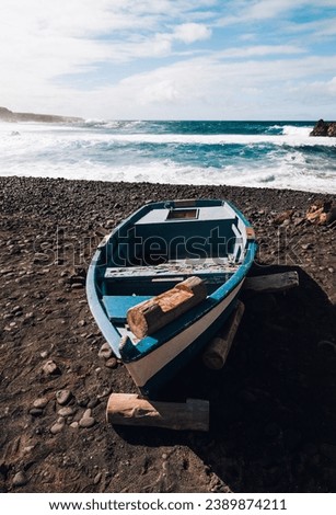 Landscape Photo of blue old boat (vessel) on volcanic black sand beach with beautiful waves in ocean on background - Lanzarote Island. Landscape shot of boat on the shore.
