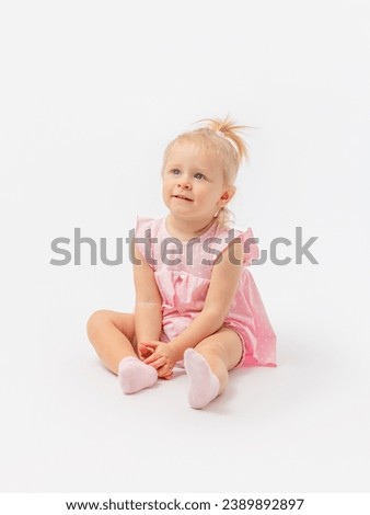 A cute blonde girl 1-2 years old in a pink dress is sitting on the floor and enthusiastically looking at something on a white background. Copy space.