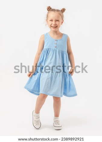 A cute Caucasian 5-year-old girl with a beautiful smile with a fashionable hairstyle stands on a white background in full height in a blue summer sundress and laughs with narrowed eyes. Royalty-Free Stock Photo #2389892921