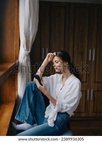 Young woman sitting at the window on a chair with a phone in hand smiling and looking out the window, spring mood, home cozy atmosphere, aesthetic lifestyle.