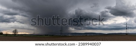 Panoramic view of a thunderstorm moving over the Taunus with grey clouds, with ploughed and fallow fields in the foreground and a high-voltage overhead power line