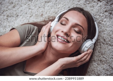 Beautiful woman in casual clothes and headphones is listening to music and smiling while lying on the floor at home