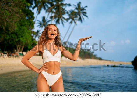 Cheerful woman in bikini presenting with hand on beach. Smiling female model showcases product placement outdoor. Summer beachwear fashion, tropical coastline lifestyle. Advertising, marketing concept