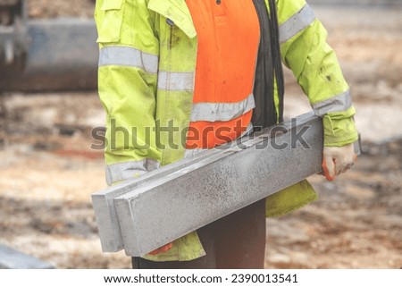 groundworker in orange and yellow hi-viz carrying heavy concrete edging curbs on the building site during new road construction. Manual handling safety concept  Royalty-Free Stock Photo #2390013541