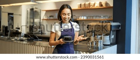 Portrait of smiling asian female barista, making coffee, holding cup of tea and taking it to cafe client, wearing apron, standing near counter.