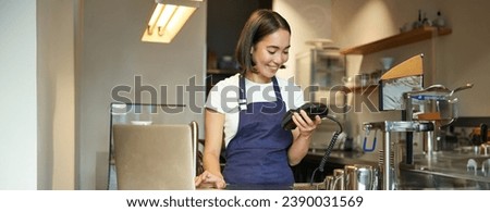 Smiling asian barista using card machine to process clients payment, guest in coffee shop paying for order.