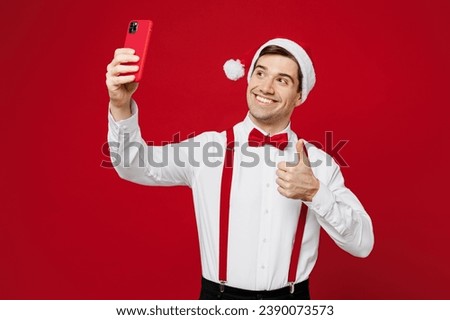 Merry young man wears white shirt Santa hat posing doing selfie shot on mobile cell phone show thumb up isolated on plain red background studio. Happy New Year Christmas celebration holiday concept