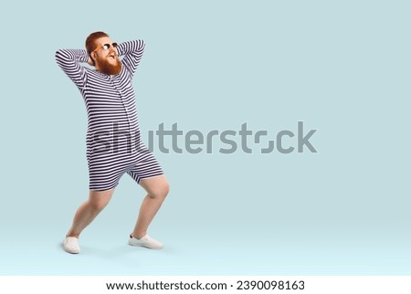 Funny fat man in swimwear having fun on the beach during summer holiday. Happy cheerful excited plump young guy wearing striped swimsuit or underwear posing on light blue copy space studio background Royalty-Free Stock Photo #2390098163