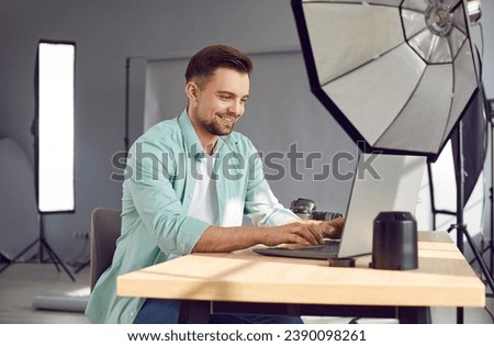 Smiling male professional photographer using laptop in photo studio. Cheerful handsome young man choosing and editing photos after photoshoot studio with professional photographic equipment