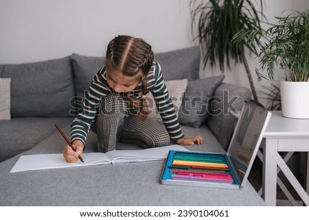 Beautiful little girl is drawing in an album while sitting on the sofa. Girl uses pencils