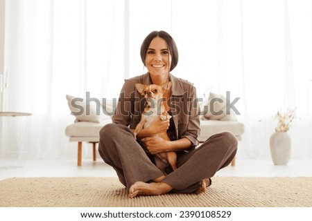 Positive brunette woman in casual clothes holding chihuahua dog in living room	