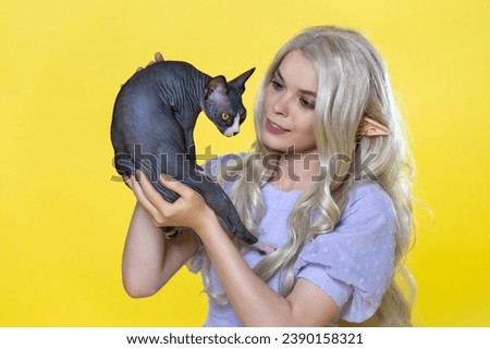 Young blonde cosplay elf in blue summer dress with beautiful curly long hair and pearls in her ear holding Sphinx kitten in hands and smiles, looking at it with tenderness. Yellow background.