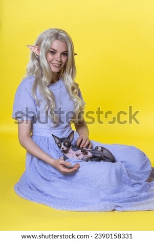 Cosplayer elf young blonde woman in blue dress sits on yellow background, holds Sphinx cat on lap, smiles and looks at camera with eyes of different colors. Elf has curly long hair and pearls in ear.