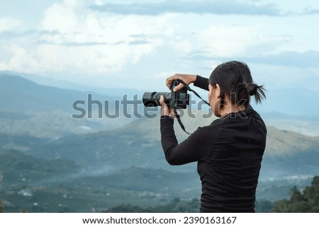 Beautiful young female photographer standing holding a camera on a mountain full of camping equipment, background view of mountains and clouds, sunset.