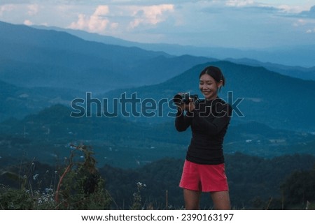Beautiful young female photographer standing holding a camera on a mountain full of camping equipment, background view of mountains and clouds, sunset.