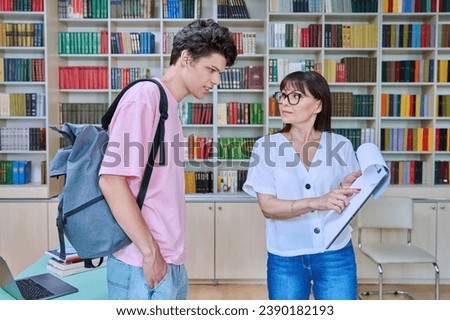 Middle-aged female teacher talking to male college student inside library