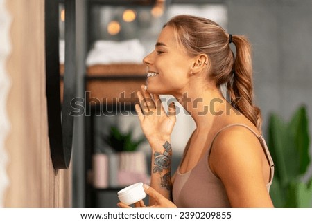 Portrait of attractive smiling woman applying face cream looking in mirror standing in modern bathroom. Skin care, cosmetology, corning routine concept 