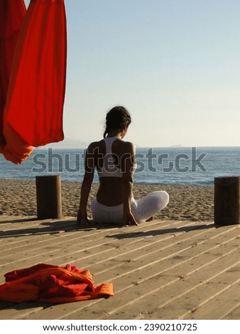 Picture of yoga asana,from the back of a brown skin woman sitting in a lotus position,dressed in white sportswear, contemplating the ocean's horizon, blue sky, red hammock,wooden deck and yellow sand