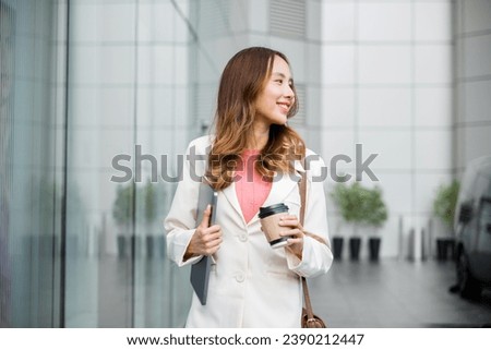 Business woman wearing white suit jacket and smile while go to work outdoor mirror building background. Confident Businesswoman with a cup of coffee and holding Laptop walking outside office building