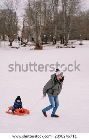 Smiling dad carries a little boy on a sled across a snowy plain at the edge of a forest