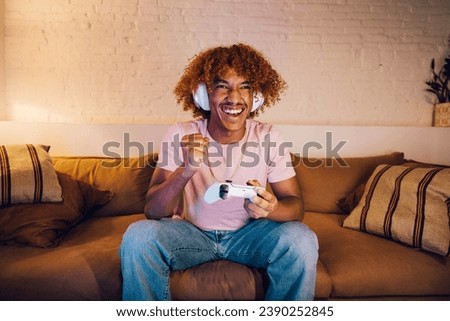 Young man with white headphones playing video games on a sofa in a living room. Happy young man celebrating a winning