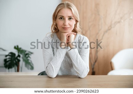 Calm female sitting behind work desk at home office looking at camera, smiling nice. Confident woman work online with clients, recording video for internet blog, provide remotely psychotherapy session Royalty-Free Stock Photo #2390262623