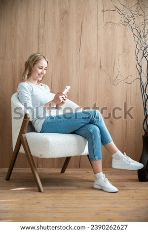 Vertical shot of smiling woman sitting on comfortable armchair and using modern smartphone. reading good news or funny messages in online app. Cozy space with wooden floor and wall in apartment Royalty-Free Stock Photo #2390262627