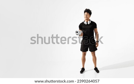 Athletic young latin man smiles with wireless headphones on neck, holding fitness mat while standing ready for gym workout on white studio backdrop. Free space for sport advertisement