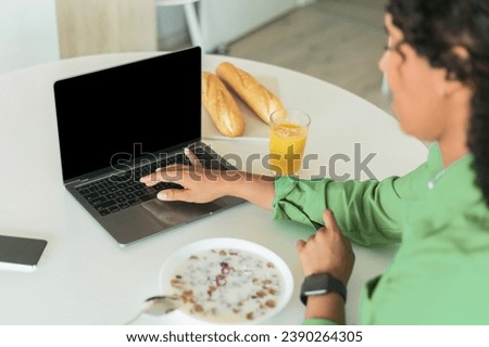 Unrecognizable black woman using laptop with blank screen while having breakfast in modern kitchen at home, cropped. Website and online offers advertisement. Mockup