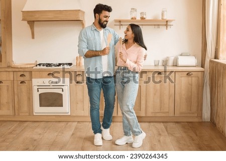 Joyful young indian couple sharing light-hearted moment while holding kitchen utensils, standing in spacious kitchen with wooden cabinets and modern design Royalty-Free Stock Photo #2390264345