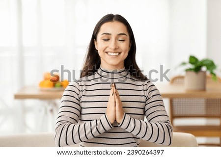 Cheerful calm young arab woman meditates with closed eyes on sofa, make pray sign in light living room interior. Free time and rest, relax, break and lifestyle cozy at home, hope, wish come true