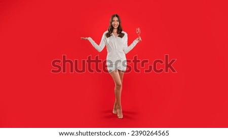 Young European woman raises her glass posing wearing elegant white dress, stands over red studio backdrop, toasting and enhancing holiday festive mood, spirit of celebration. Panorama