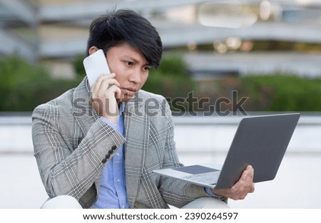 Displeased Korean manager man using laptop, talking with phone in hand, confronts digital challenges while working online outdoors, reflecting on business strategies outside bustling city office