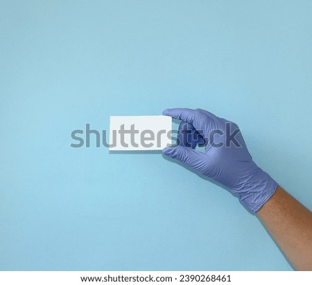 A doctor's hand in a sterile medical glove holds a blank sheet of paper on a blue background. Blank business card for text. Close-up.